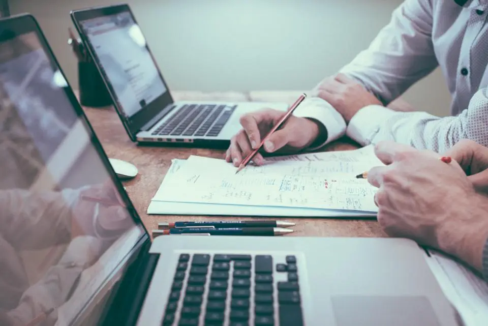 Dos personas revisando notas y esquemas sobre una mesa con portátiles